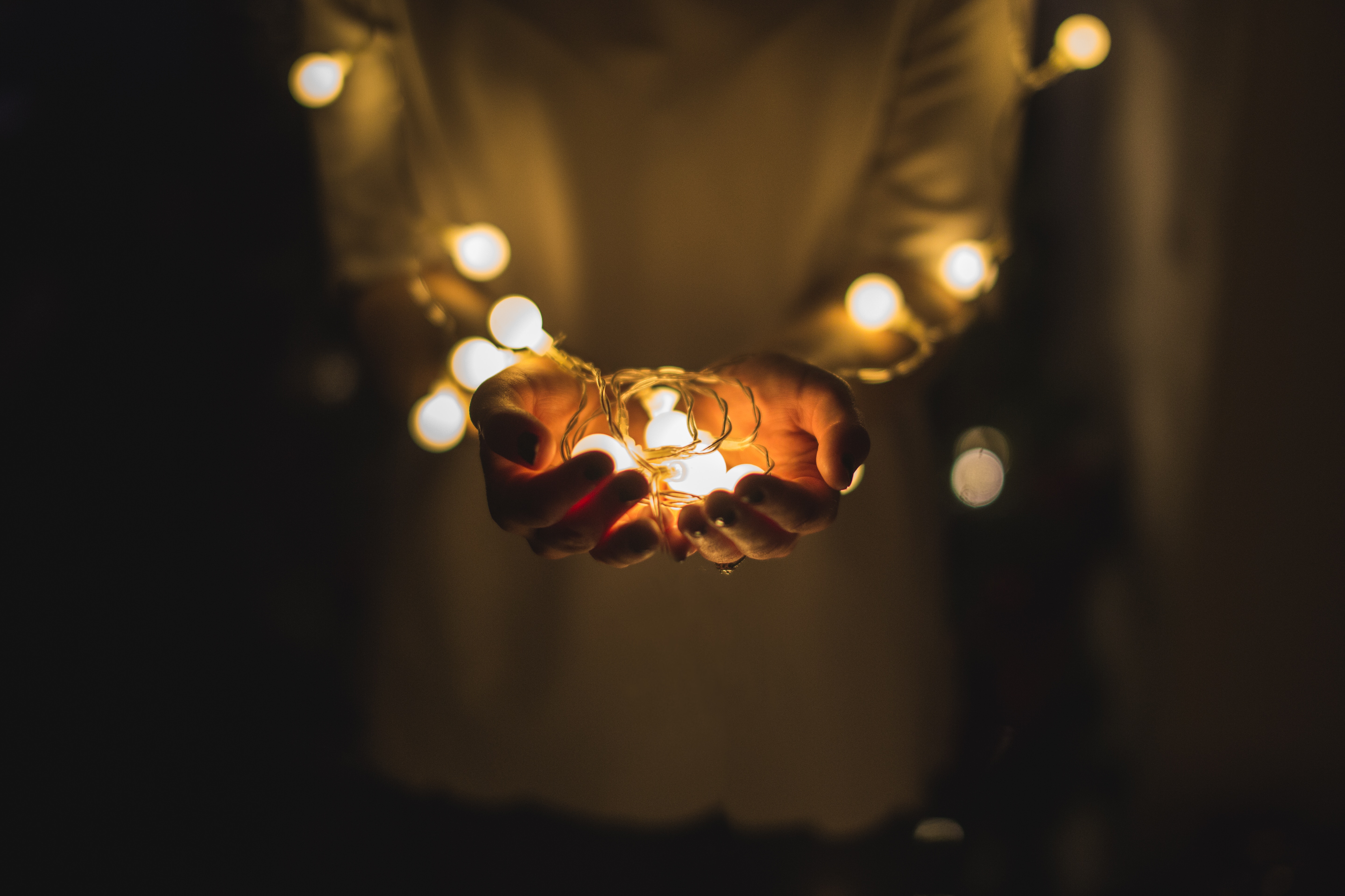 Arms, hands and torso of person in white shirt. Arms wrapped in a string of white lights. Lights are pooled into hands, and the hands are cupped in front and held out as if in offering.